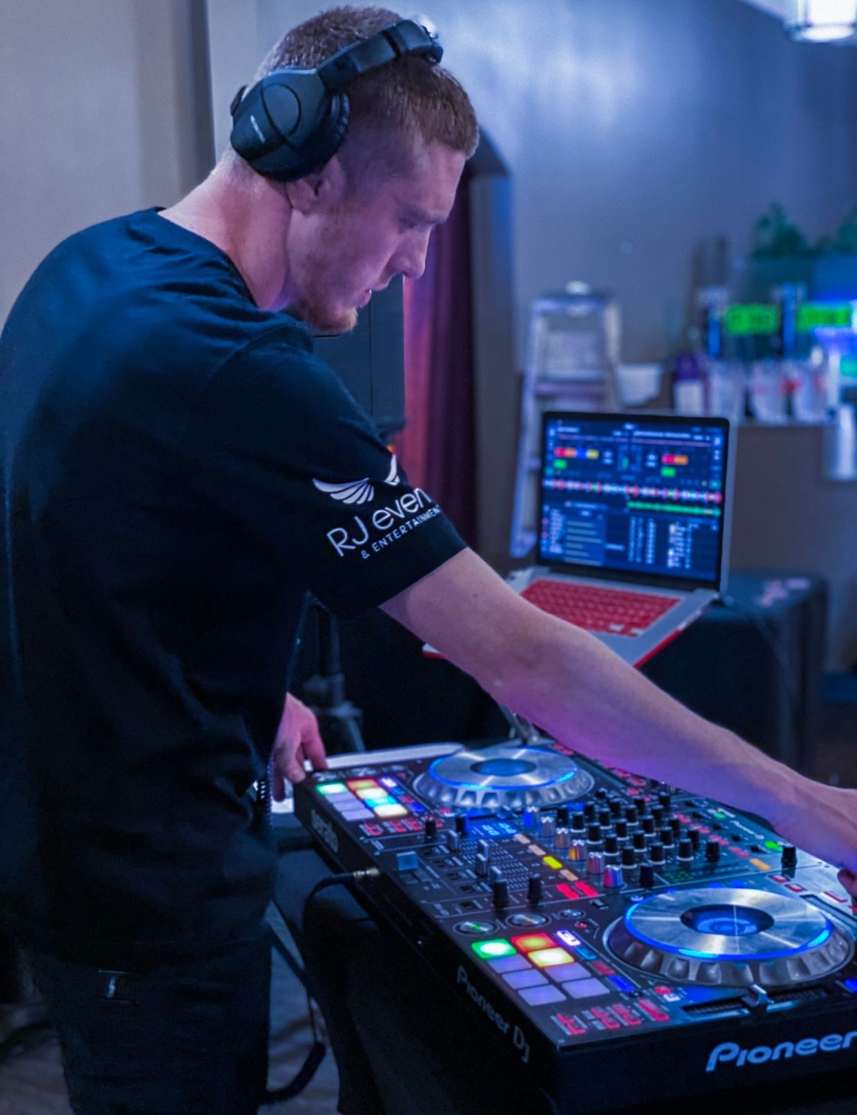 DJ wearing headphones and operating a Pioneer DJ controller with a laptop showing music software in the background.