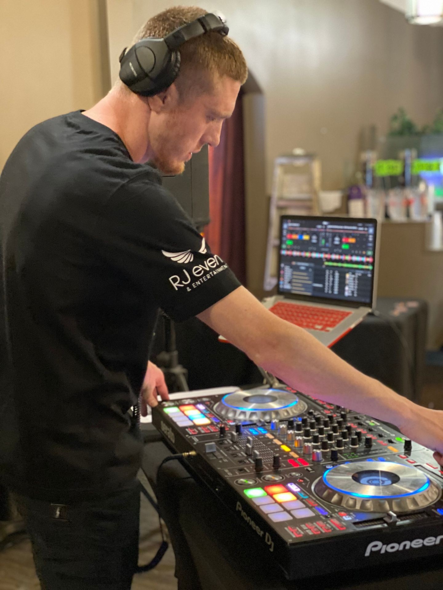 DJ wearing headphones, mixing on a Pioneer DJ controller with a laptop in the background displaying software.